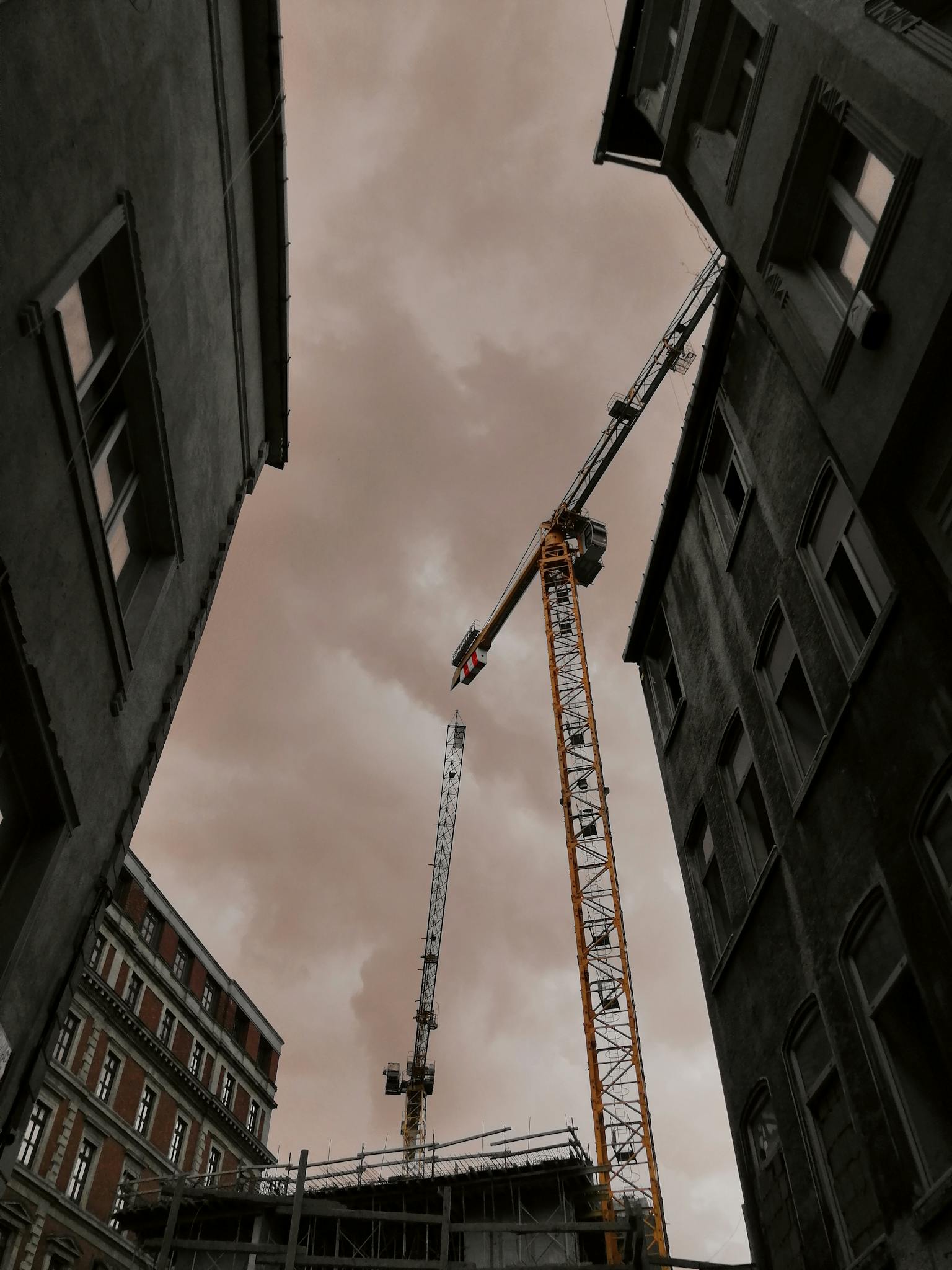 Dramatic view of construction cranes between buildings under a cloudy sky framed by urban architecture.