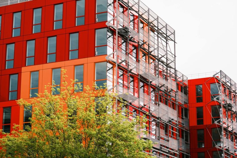 Striking red building facade with windows and scaffolding surrounded by greenery captures urban development.